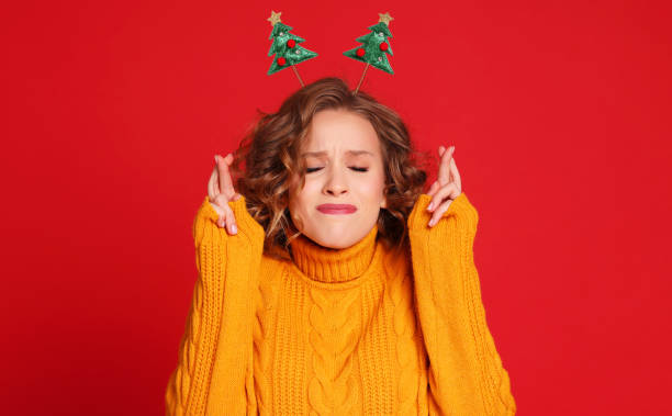 la femme gaie avec le bandeau de noël croise des doigts pour la bonne chance, prie et espère que les rêves se réalisent, dans la nouvelle année, attend pour le miracle ferme les yeux sur le fond rouge - faire un voeu photos et images de collection