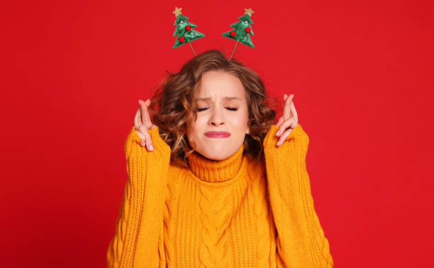 크리스마스 머리띠를 가진 쾌활한 여자는 행운을 위해 손가락을 교차하고, 기도하고 꿈이 실현되기를 희망하며, 새해에는 붉은 배경에 대한 기적의 눈을 닫습니다. - 소원 빌기 뉴스 사진 이미지