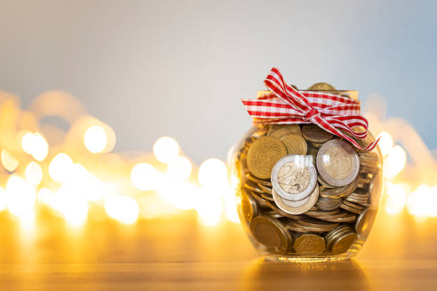 barattolo di risparmio pieno di monete davanti alle luci di natale - jar coin currency glass foto e immagini stock