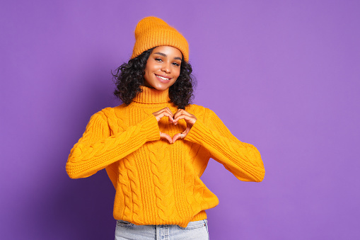 cheerful ethnic African-American young  woman in yellow sweater and hat shape hearts to camera and express love and gratitude,   confess in good feelings on colorful  purple background
