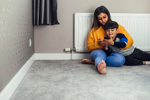 A shot of a mid adult woman and her little boy wearing casual clothing. They are sitting on the floor by a radiator and are controlling the heating with a home control centre on a mobile phone.