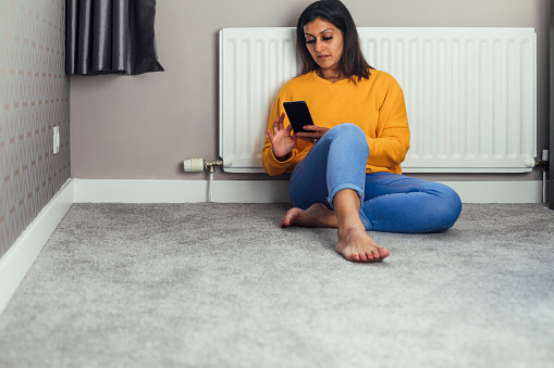 A shot of a mid adult Pakistani woman wearing casual clothing in her home. She is sitting on the floor next to a radiator and using a home control centre on her phone to turn the heating on.
