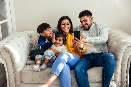 A shot of a mid adult couple and their two young sons wearing casual clothing. They are all sitting together on a sofa in their living room on a video call.
