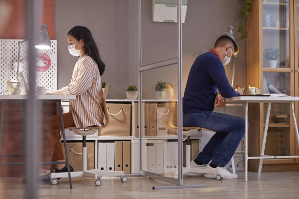 Two Office Workers in Cubicles Side view portrait of two people wearing masks in office while working at desks in separate cubicles post pandemic, copy space office cubicle mask stock pictures, royalty-free photos & images