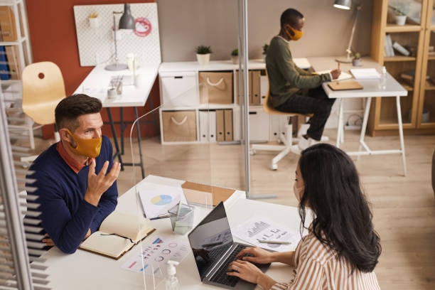 Post Pandemic Business Meeting High angle portrait of modern mature man wearing mask and talking to colleague through glass panel during post pandemic business meeting, copy space office cubicle mask stock pictures, royalty-free photos & images