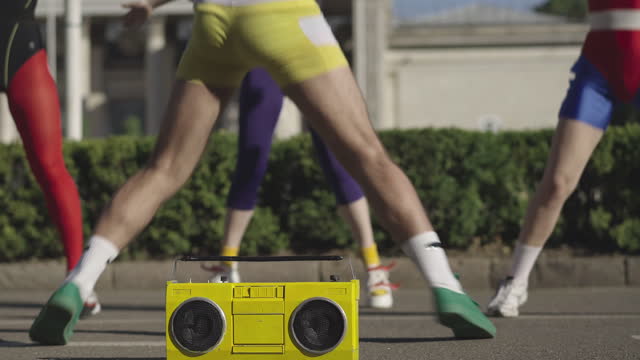 Unrecognizable group of fit Caucasian man and women jumping at the background to disco music from yellow vintage tape recorder standing on road at front. 1980s sportive lifestyle concept.