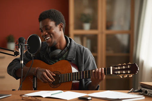 Portrait of talented African-American man singing to microphone and playing guitar while recording music in studio, copy space