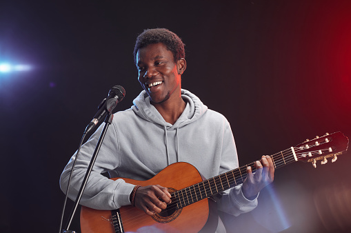 Waist up portrait of young African-American man playing guitar on stage and singing to microphone in lights, copy space