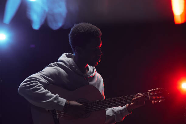 African-American Musician Playing Acoustic Guitar on Stage Side view outline of young African-American man playing acoustic guitar on stage in colored lights, copy space acoustic music photos stock pictures, royalty-free photos & images