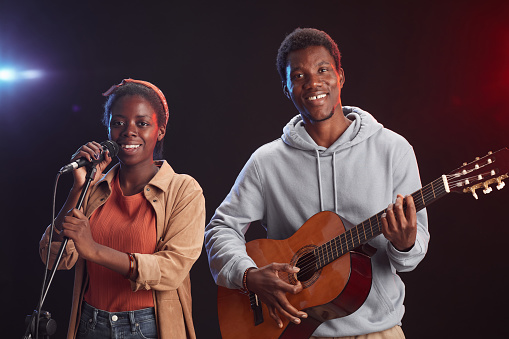 Waist up portrait of two African-American musicians playing guitar on stage and singing to microphone smiling