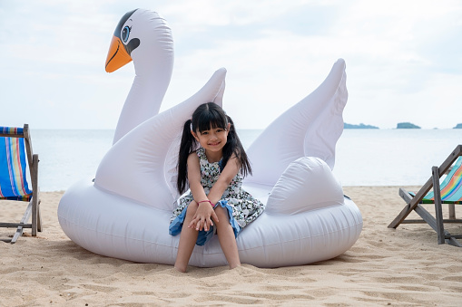 Happy Asian girl kid sitting on swan Floating raft on sand beach summer vacation