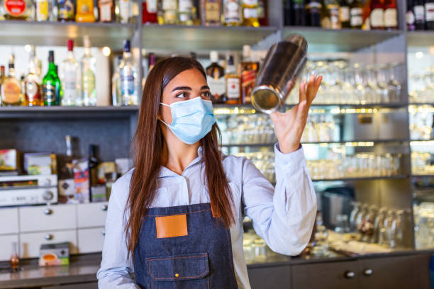 istock Young female worker at bartender desk in restaurant bar preparing coctail with shaker. Beautiful young woman behind bar wearing protective face mask 1287425917