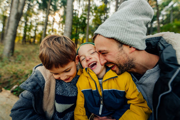 Mother and two sons are running along a narrow concrete slab path surrounded by lush green forest. The woman is in the middle, wearing a purple jacket and purple rain boots. She is holding hands with the boys dressed jackets and jeans.\nShot with Nikon D810