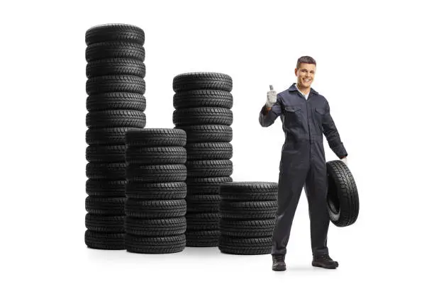 Auto mechanic with a car tire and piles of tires behind isolated on white background