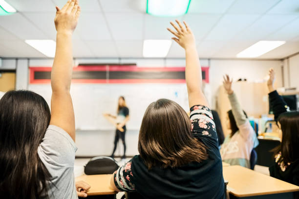 Getting the best education Rear view of a group of students sitting in classroom raising hands to answer the questions hand raised classroom student high school student stock pictures, royalty-free photos & images