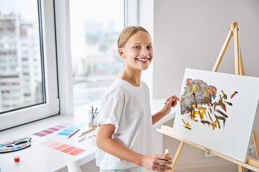 Smiling happy beautiful blonde beginner painter standing by the easel in her home art studio