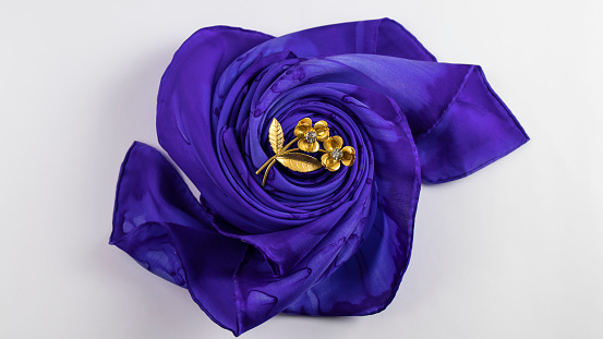 Purple silk scarf folded in the shape of a flower and gold vintage brooch with sparkling crystals on the white background. Isolated object.