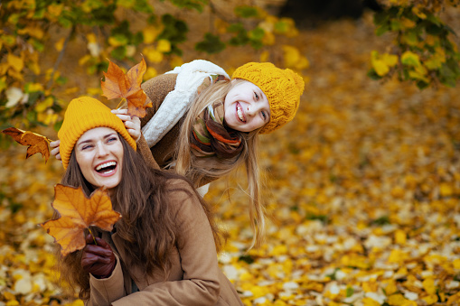 Hello october. happy young mother and daughter in orange hats with autumn yellow leaves having fun time outside in the city park in autumn.