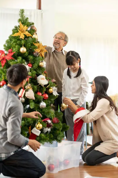 Multigenerational asian Family decorating a Christmas tree. Mom Dad daughter girl and grandfather decorate the Christmas tree prepare for season greeting of Merry Christmas and Happy Holidays.
