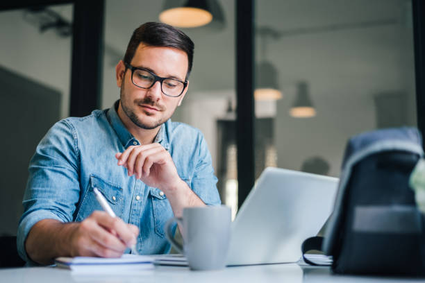 istock Close up portrait of handsome man working from home office taking reading and writing notes in note pad while working on laptop computer casual businessman entrepreneur focused doing research 1287455348