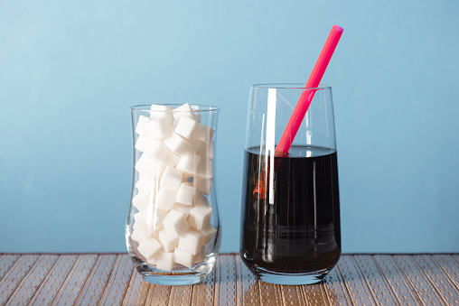 Two drink glasses in front of blue background.  The first glass is filled with sugar cubes and the second glass is filled with cola and has a red pipette in it.