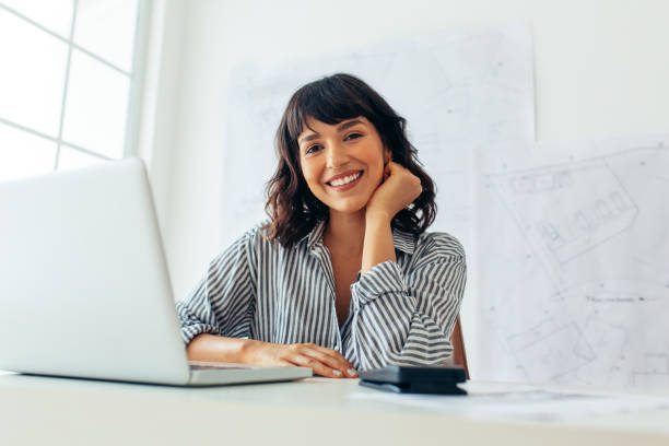 smiling female architect sitting at her office desk - trabalhador imagens e fotografias de stock