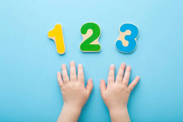 Photo of Baby hands and colorful wooden numbers 123 on light blue table background. Pastel color. Closeup. Point of view shot. Time to learning. Top down view.