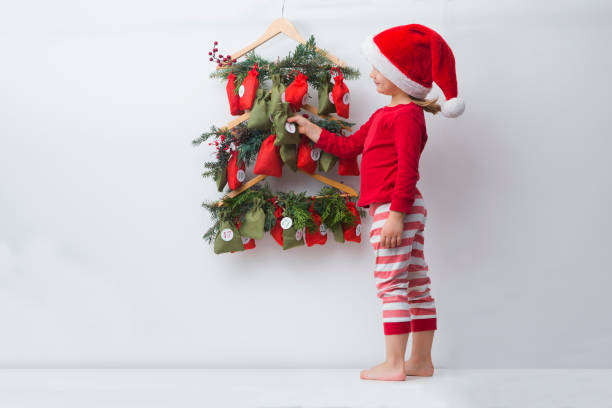 una niña con sombrero de navidad y pijama en busca de dulces y regalos en el calendario de adviento. - advent calendar advent christmas childhood fotografías e imágenes de stock