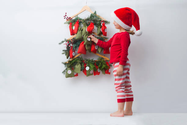 a child girl in a christmas hat and pajamas searching for candy and gifts in advent calendar. - advent calendar advent christmas childhood imagens e fotografias de stock