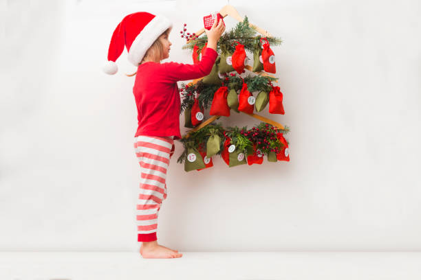 una niña con sombrero de navidad y pijama en busca de dulces y regalos en el calendario de adviento. - advent calendar advent christmas childhood fotografías e imágenes de stock