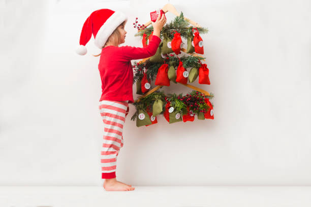 a child girl in a christmas hat and pajamas searching for candy and gifts in advent calendar. - advent calendar advent christmas childhood imagens e fotografias de stock