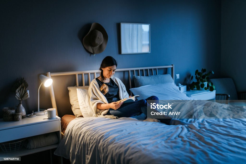 Woman reading a book at night Young woman lying on the bed and reading a book at night. The room is dark and blue and the light on the night stand is illuminating the book. Bedroom, horizontal photo Reading Stock Photo