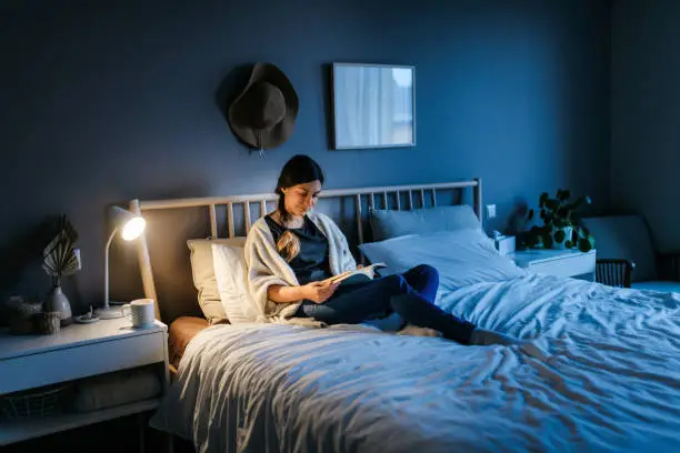 Young woman lying on the bed and reading a book at night. The room is dark and blue and the light on the night stand is illuminating the book. Bedroom, horizontal photo