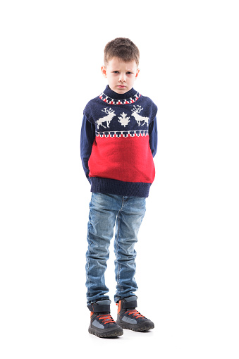 Angry cute young boy child in Christmas sweater looking at camera frustrated with hands behind. Full body portrait isolated on white background.