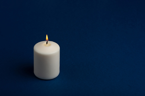 One white burning candle on dark blue background with copy space for text. Close up.