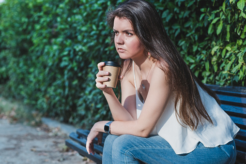portrait of a young caucasian woman dressing casual with jeans and a white shirt and sitting on a bench of a park with a cup of a paper take away coffee. Selective focus on paper cup