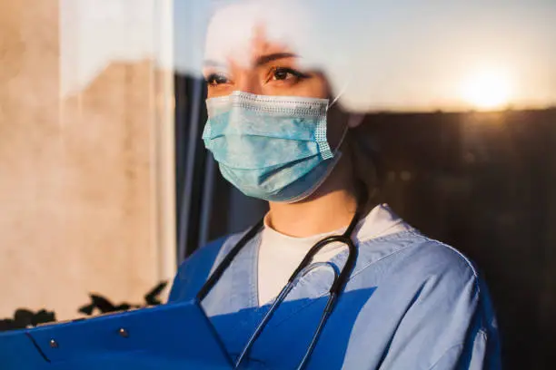 Young sad female caucasian NHS EMS doctor carer looking through ICU window, fear uncertainty in eyes, wearing face mask gazing at sun ,hope and faith in overcoming Coronavirus COVID-19 pandemic crisis