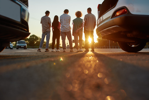 Hanging out. Backs of five young ordinary dressed friends standing next to each other outside on a parking site with their cars on a front line casting shadows on a camera during a beautiful sunset