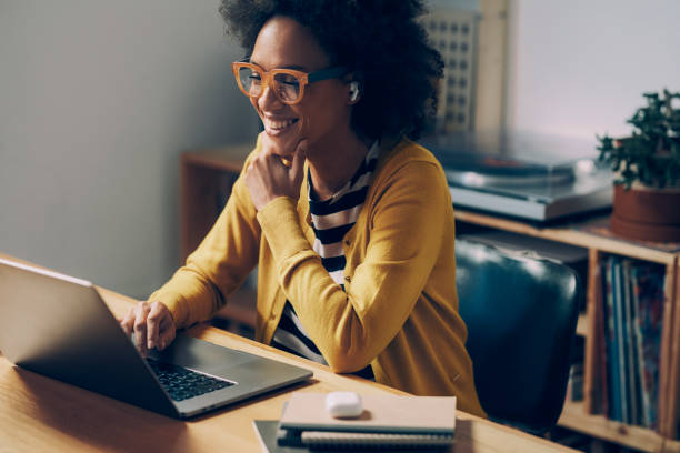 smiling african american woman wearing glasses and wireless earphones makes a video call on her laptop computer at her home office - desk meeting conference business imagens e fotografias de stock