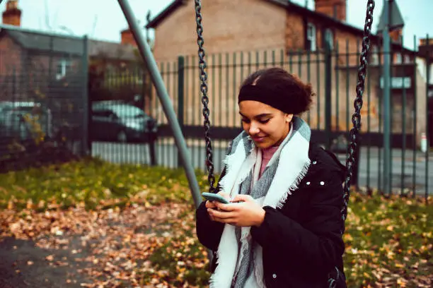 Stock photo of a teenager girl (14-15) who is texting on her mobile phone while sitting on swing in the playground. She is relaxed and smiling. She is wearing winter clothing. This file has a signed model release.