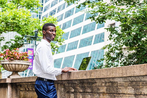 Young African American College Student studying in New York City, wearing white long sleeve shirt, blue pants, standing on campus, working on laptop computer on top of old style fence wall, thinking.