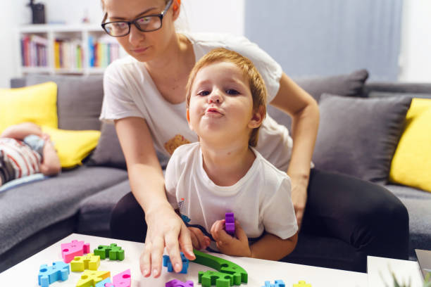 istock Front view portrait of small caucasian boy looking to the camera while playing with his mother by the table on the sofa couch at home - growing up family bonding leisure concept 1287503210