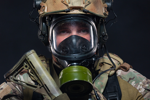 Close up portrait of the face and tired eyes of a soldier standing in a military helmet and gas mask, ready for a chemical attack, isolated on a black background.