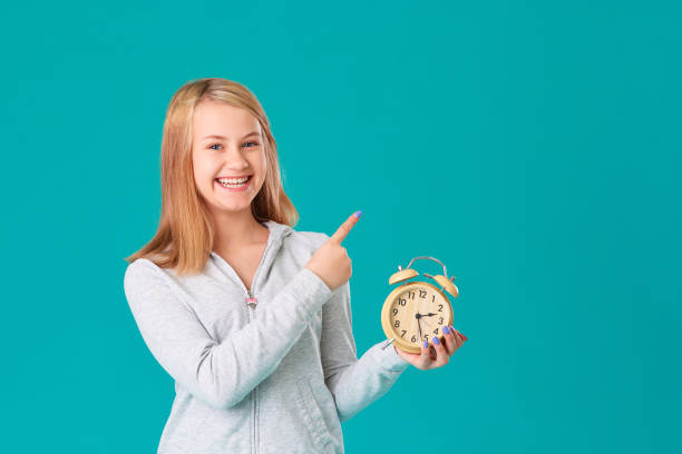 한 십대 소녀가 고립된 청록색 배경에 서서 행복하고 웃으며 알람 시계를 들고 있습니다. 텍스트의 빈 공간에 손 제스처를 만듭니다. - 12 oclock 뉴스 사진 이미지