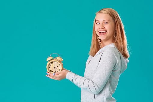 A teenage girl holds an alarm clock, standing on an isolated turquoise background, very happy and excited, the expression of a winner , she celebrates the victory by shouting with a big smile.