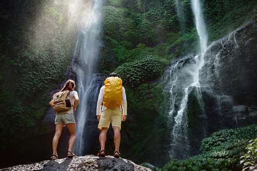 Couple of travelers or hikers with backpacks are standing on big rock and enjoying big waterfall