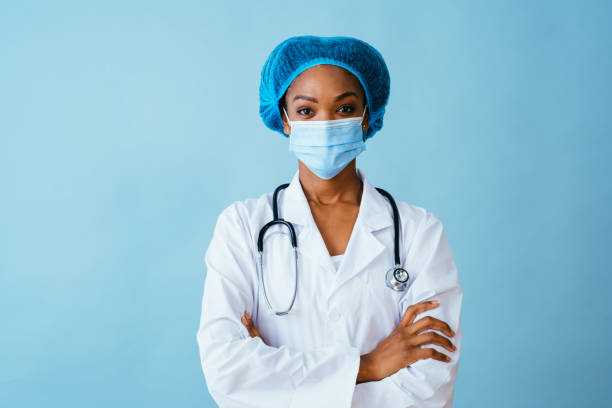 porträt eines arztes im labormantel mit gesichtsmaske und haarschutz mit gekreuzten armen, isoliert auf blauem hintergrund - lab coat women one person female stock-fotos und bilder