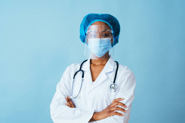 portrait of a doctor in lab coat with face mask, face shield,  and hair ppe with arms crossed - stethoscope blue healthcare and medicine occupation imagens e fotografias de stock