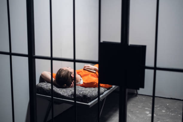 istock Lights out in prison. A convict in an orange robe rests on a bunk in a cell with a book 1287563221