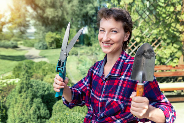 retrato de jovem adulto feliz linda morena branca trabalhadora cultivadora segurando tesouras e pá de exibição com jardim de flores verde formal ao fundo. jardineiro sorridente no quintal de casa - gardening women vegetable formal garden - fotografias e filmes do acervo